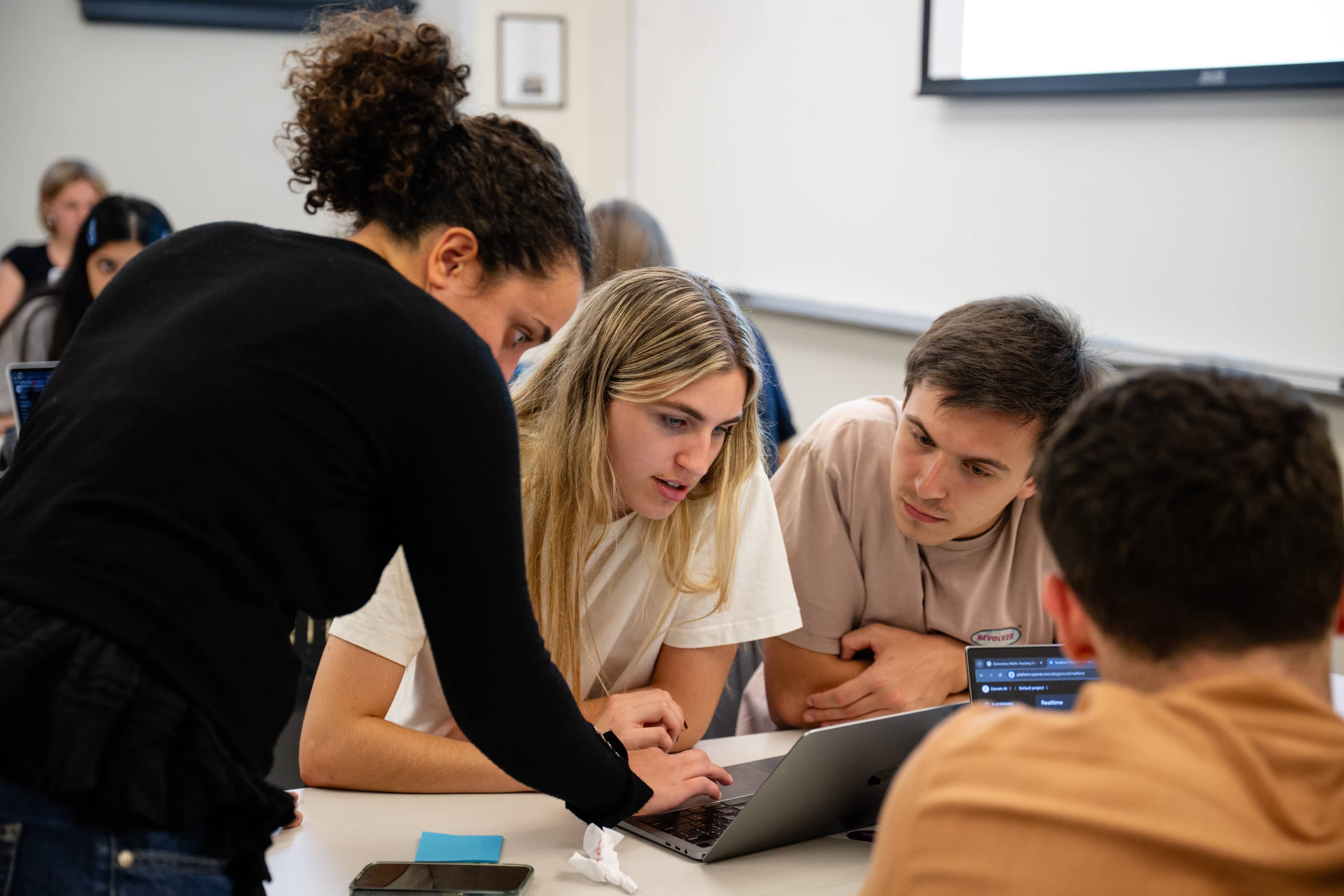 Students seated at a table receive help from a standing instructor who leans over their work.