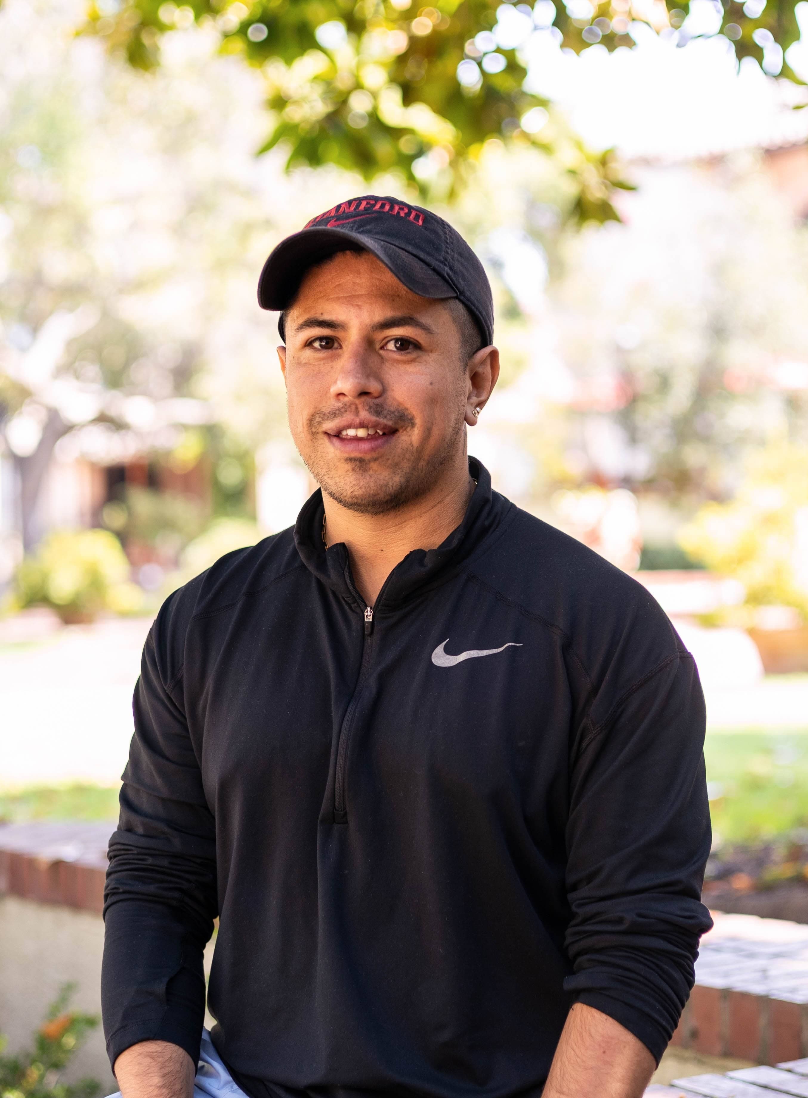 Jeiti wears a black shirt and a hat and stands outside under a tree.