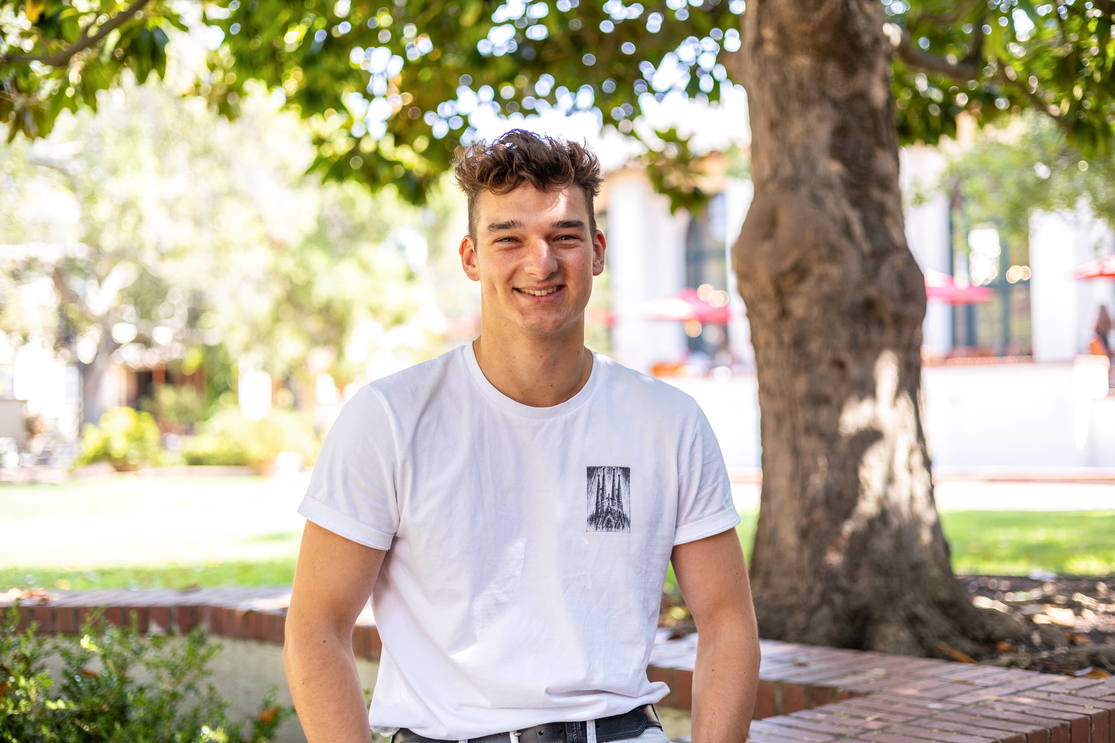 Max sits under a tree and wears a white shirt.